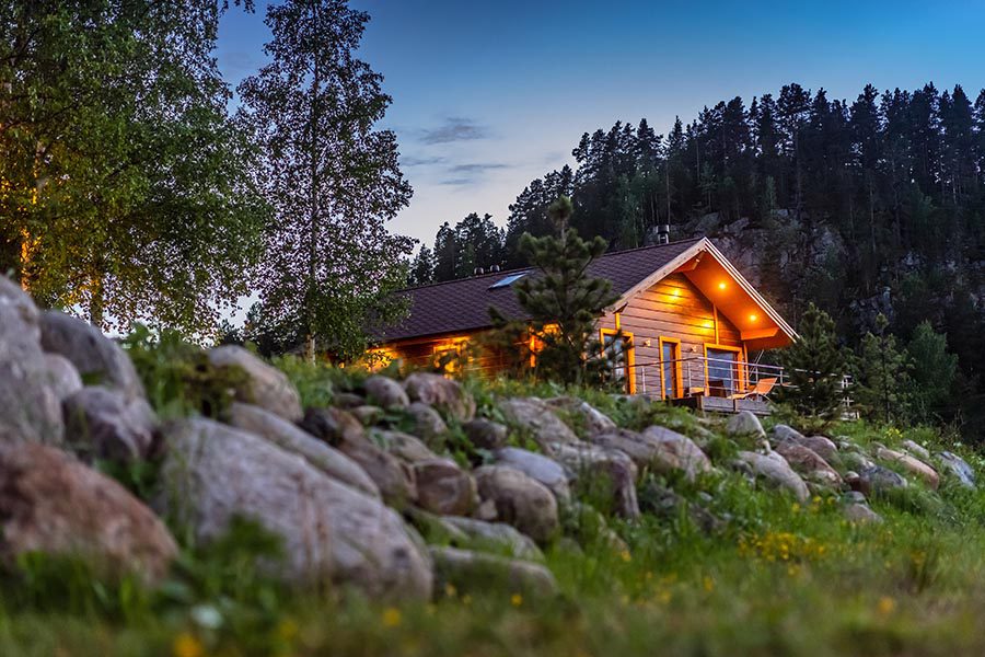 Brush Zone Homeowners Insurance - Beautiful Wood Home Lit Up for the Evening, Surrounded by Large Boulders, Trees and Grass, A Large Tree-Covered Hill Behind the Home