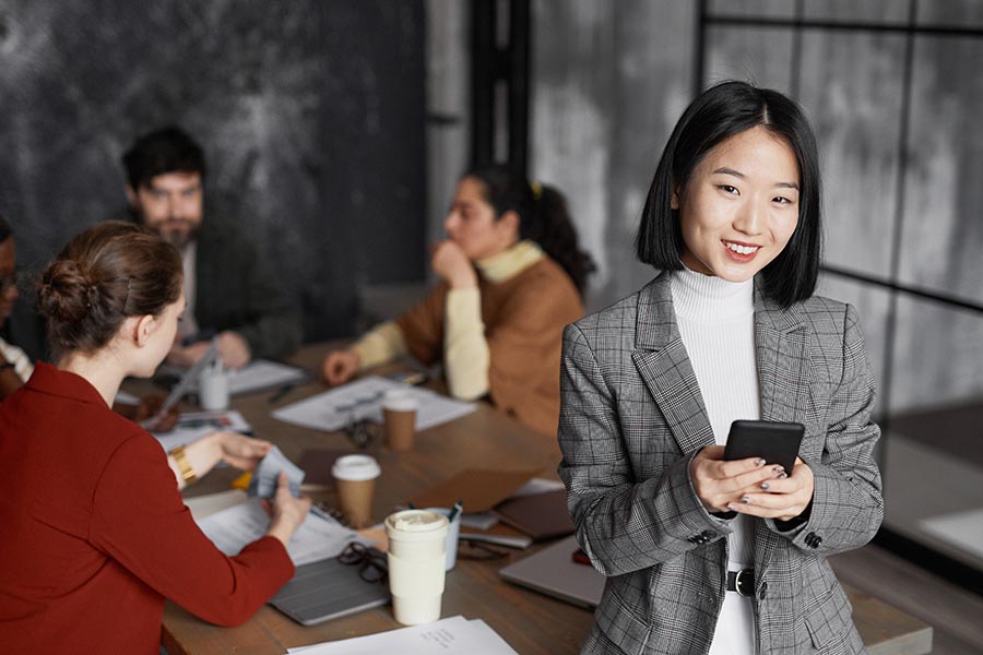 Employee Benefits - Boss Holding a Phone Standing in Front of a Lively Meeting At a Conference Table